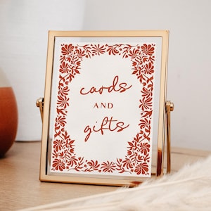 May include: A gold-framed sign with the words "cards and gifts" in a handwritten font. The sign is surrounded by a floral border in a reddish-orange color. The sign is displayed on a light-colored surface.