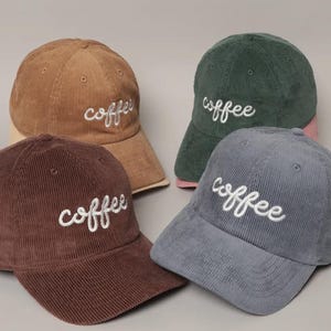 Puede incluir: Cuatro gorras de béisbol de pana en tonos marrón, verde y azul. Cada gorra tiene la palabra "coffee" bordada en escritura blanca. Las gorras tienen un diseño clásico de seis paneles con una visera curva.