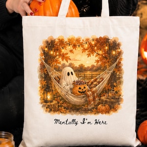 May include: A cream-coloured canvas tote bag featuring a printed illustration of a ghost relaxing in a hammock between two trees, holding a jack-o'-lantern bucket filled with sweets. The text "Mentally I'm Here" is printed below the graphic.
