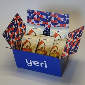 Puede incluir: Una caja azul con la palabra "yeri" en blanco en la parte delantera. Contiene paquetes blancos con la palabra "yeri" y una ilustración. El interior y las solapas de la caja presentan un patrón geométrico rojo, blanco y azul.