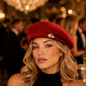 May include: A deep red felt beret featuring a gold-toned decorative pin with a central white pearl accent. The beret has a smooth, rounded crown and a structured brim, providing a classic silhouette that sits comfortably on the head.