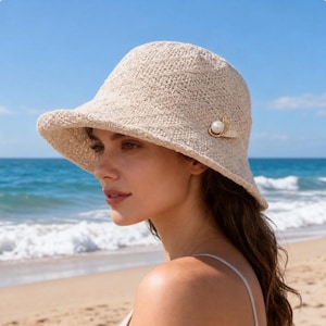 May include: A beige woven bucket hat featuring a textured, open-knit pattern. The hat is adorned with a decorative gold-tone metal accent piece on the side, which is set with a single round white pearl and small clear crystals.