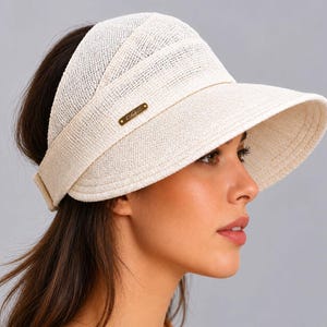 May include: A cream-colored straw visor featuring a wide, curved brim and a textured, woven fabric construction. The visor includes a small, rectangular gold-toned metal plaque attached to the side, displaying engraved text branding.