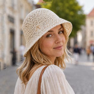 Może przedstawiać: Kremowy szydełkowy kapelusz typu bucket hat o ażurowej fakturze z małą, wyhaftowaną złotą literą M z boku. Kapelusz posiada zaokrągloną główkę i opadające rondo, wykonany jest z lekkiego, plecionego materiału o wyraźnym splocie.