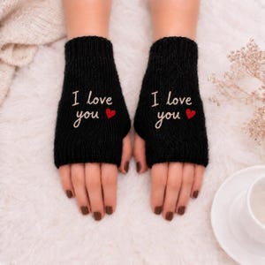 Może przedstawiać: Para czarnych rękawiczek bez palców z wyhaftowanym białym napisem "I love you" oraz małym czerwonym sercem na wierzchu każdej dłoni. Rękawiczki wykonane są z dzianiny o prążkowanej fakturze i zakrywają dłoń, pozostawiając palce odsłonięte.