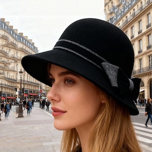 May include: A black cloche hat with a wide brim and a gray ribbon detail. The hat features a bow on the side. The hat is made of a soft material and is suitable for various occasions.