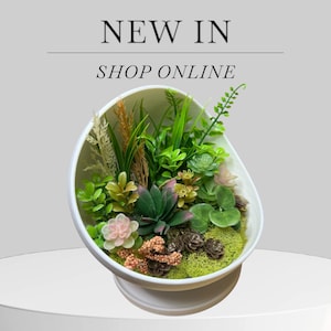 Puede incluir: Una maceta blanca en forma de huevo llena de suculentas y vegetación artificiales. El arreglo incluye plantas verdes, rosas y amarillas, pequeñas piñas y musgo. El texto "NEW IN" y "SHOP ONLINE" se muestra encima de la maceta.
