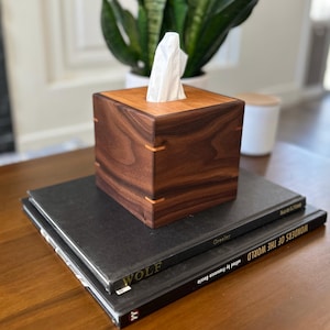 May include: A square tissue box cover crafted from dark brown walnut wood with a lighter wood top panel. The box features visible light-colored wood splines at the corners and holds a single white tissue extending from the center opening.