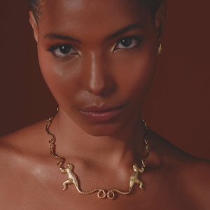 May include: A gold necklace with two monkey pendants. The necklace is worn around the neck and is set against a brown background. The monkey figures are connected by a chain with a textured surface.