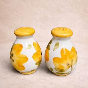 May include: A pair of ceramic salt and pepper shakers. The shakers are white with yellow floral designs and yellow lids. The floral design includes yellow flowers and green leaves. The shakers are on a neutral background.