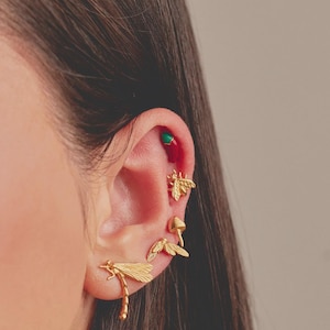 Peut inclure: Gros plan sur une oreille ornée de boucles d'oreilles dorées. Les boucles d'oreilles comprennent une libellule, une abeille, un champignon et une paire de boucles d'oreilles ailées. Une petite boucle d'oreille verte et rouge est également visible.