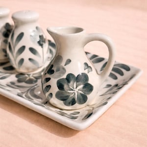 May include: A white ceramic serving set with a floral design in shades of gray. The set includes a small pitcher with a handle, two salt shakers, and a rectangular tray. The floral pattern is repeated on each piece.