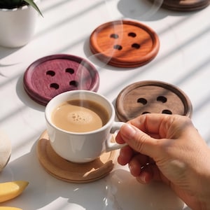 May include: A set of circular wooden coasters shaped like four-hole buttons in various wood tones including light, dark, reddish, and purple hues. A white ceramic mug filled with a steaming beige beverage rests on the light-colored wooden coaster.