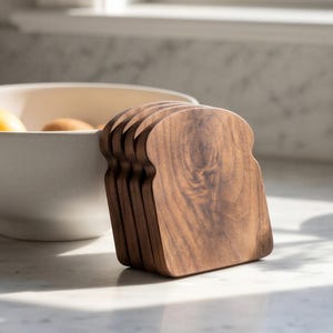 May include: A stack of five wooden bread-shaped cutting boards. The boards are made of a dark brown wood with visible grain patterns. They are arranged vertically on a white marble countertop, near a white bowl.