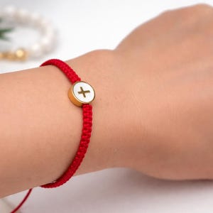Könnte beinhalten: Rotes geflochtenes Armband mit einem goldfarbenen, kreisförmigen Anhänger mit einem weißen Kreuz. Das Armband wird am Handgelenk getragen. Im Hintergrund ist ein weißes und goldenes Perlenarmband zu sehen.