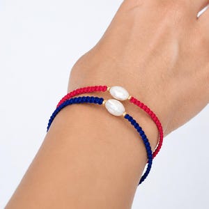 May include: Two braided cord bracelets, one in vibrant red and one in deep navy blue, each featuring a single oval-shaped white freshwater pearl centred between two small gold-toned metal beads on the cord.