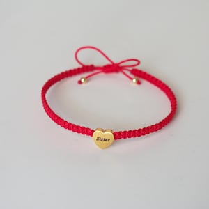 Op de afbeelding: Rode gevlochten armband met een gouden hartvormige bedel met de tekst "Sister". De armband heeft een striksluiting. De armband ligt op een witte achtergrond.