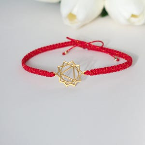 May include: Red macrame bracelet with a gold-colored charm. The bracelet features a woven design and a central charm with a geometric shape. The bracelet is displayed on a white surface with white flowers in the background.