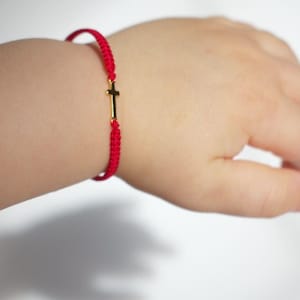 May include: A red braided bracelet with a gold cross charm is worn on a wrist. The bracelet is made of a thin, woven material. The gold cross is small and centered on the bracelet. The background is a soft, neutral color.