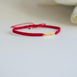 May include: Red macrame bracelet with a gold-colored bar that spells out "mama". The bracelet is adjustable and sits on a white surface. A white flower is in the background.