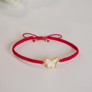 May include: Red braided bracelet featuring a gold-colored butterfly charm with a white center. The bracelet is tied with a red string and small gold-colored beads. A delicate piece of jewelry.