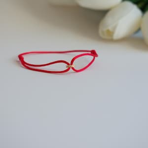 May include: A red bracelet with a gold-colored accent. The bracelet is made of a thin, flexible material and features a simple, elegant design. White tulips are in the background.