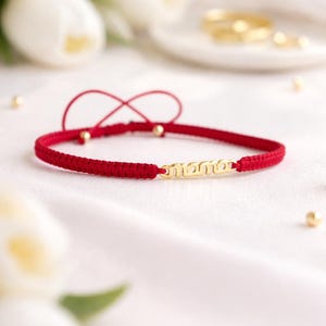 May include: A red woven bracelet with a gold-colored 'mama' charm. The bracelet features a sliding knot closure and small gold beads. The background includes white flowers and gold rings.