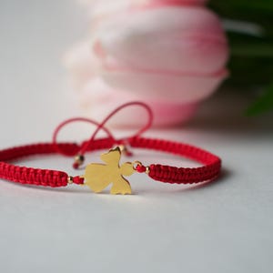May include: A red braided bracelet with a gold-colored charm in the shape of an angel. The bracelet has adjustable sliding knots and small gold-colored beads. The background features soft pink flowers.