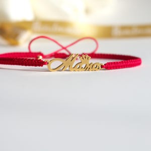 May include: A red braided bracelet with a gold-colored charm that spells "Mama" with a crown above the first "a". The bracelet is on a white surface.