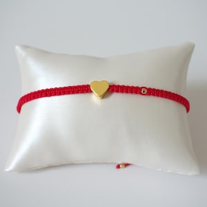 May include: A red macrame bracelet with a gold heart charm and a small gold bead. The bracelet is displayed on a white satin pillow against a white background. The bracelet is a delicate piece of jewelry.