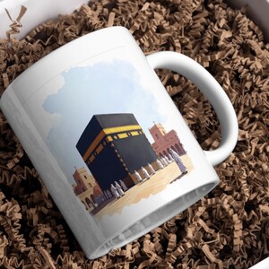 May include: White ceramic mug featuring an illustration of the Kaaba in Mecca, with a black and gold design. The mug also depicts surrounding buildings and figures, set against a blue sky background.