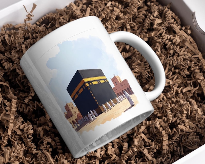 May include: White ceramic mug featuring an illustration of the Kaaba in Mecca, with a black and gold design. The mug also depicts surrounding buildings and figures, set against a blue sky background.