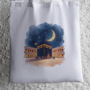May include: White tote bag with a watercolour illustration of the Kaaba at night. The artwork shows the Kaaba with a crescent moon and stars in a dark blue sky, surrounded by buildings with arched windows.
