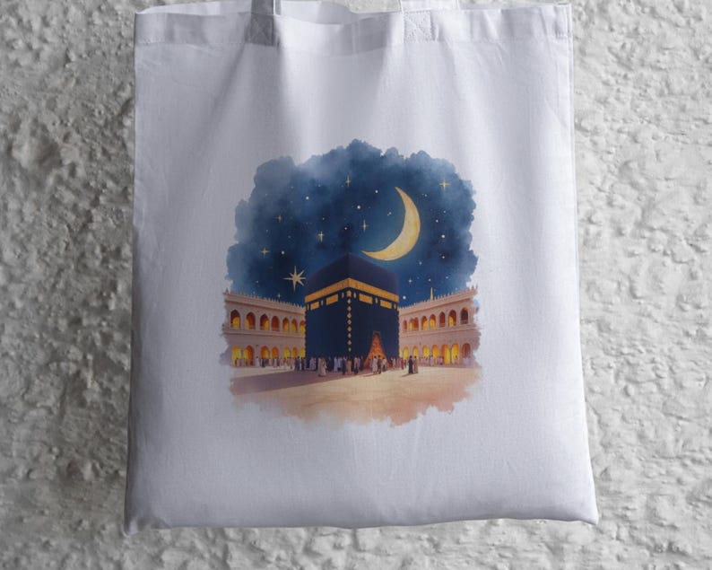 May include: White tote bag with a watercolour illustration of the Kaaba at night. The artwork shows the Kaaba with a crescent moon and stars in a dark blue sky, surrounded by buildings with arched windows.