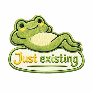 May include: Embroidered patch showing a green frog with yellow details, lounging on a white oval. The patch has the text "Just existing" in yellow and green, with a green border. The frog has black eyes and a smiling face.