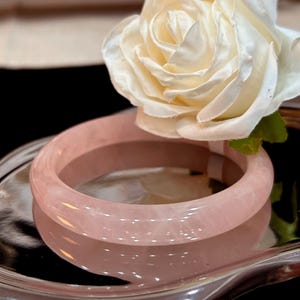 Könnte beinhalten: Ein poliertes, hellrosa Rosenquarz-Armreif-Armband. Das Armband ist glatt und abgerundet und reflektiert das Licht. Eine weiße und cremefarbene Rose liegt über dem Armband und verleiht ihm Eleganz. Das Armband wird auf einer reflektierenden Oberfläche präsentiert.
