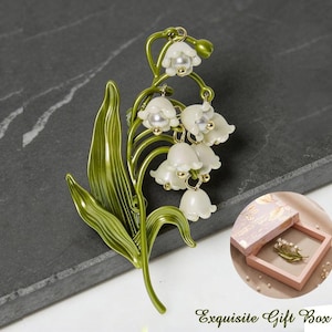 May include: A green and gold-toned metal brooch shaped like a lily of the valley plant. The brooch has white flower pendants with pearl accents. The image also includes a gift box with the text "Exquisite Gift Box".