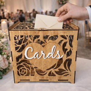 May include: A wooden card box with intricate floral cutouts and a heart-shaped design. The word "Cards" is displayed in white script. An envelope is being placed into the box, suggesting a wedding or event setting.