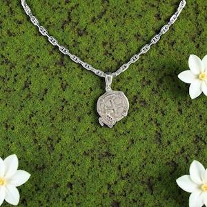 May include: A silver-toned necklace with a round pendant featuring a detailed design. The pendant is attached to a chain, set against a green moss background with white flowers. The necklace is a jewelry item.
