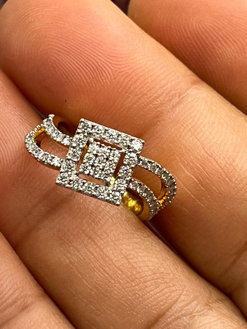 Puede incluir: Un anillo de oro con un centro cuadrado con diamantes y un dise&ntilde;o de doble banda. El anillo presenta un patr&oacute;n geom&eacute;trico con numerosos diamantes peque&ntilde;os y brillantes, creando una apariencia lujosa y elegante. El anillo se muestra en primer plano.