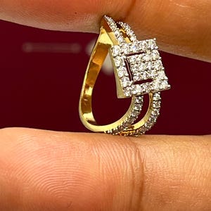 Puede incluir: Un anillo dorado con un dise&ntilde;o en forma de l&aacute;grima y un cuadrado de diamantes en el centro. El anillo est&aacute; adornado con peque&ntilde;os diamantes transparentes en la banda y alrededor del cuadrado. El anillo se sostiene entre los dedos sobre un fondo rojo oscuro.