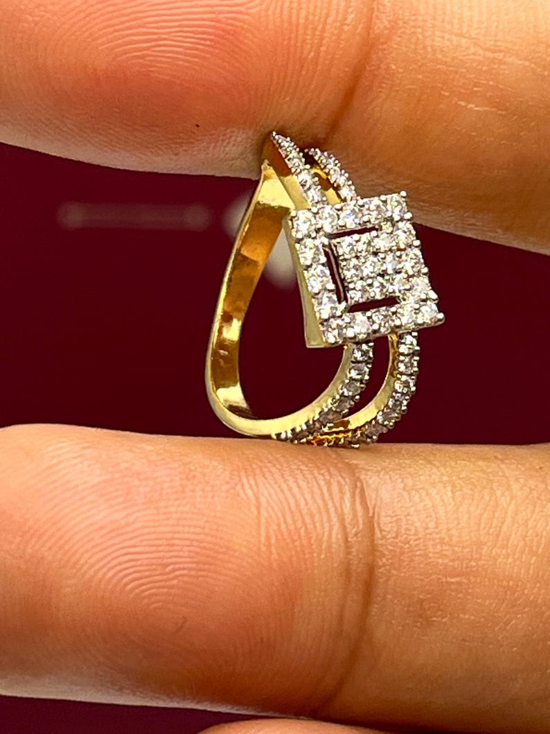 Puede incluir: Un anillo dorado con un dise&ntilde;o en forma de l&aacute;grima y un cuadrado de diamantes en el centro. El anillo est&aacute; adornado con peque&ntilde;os diamantes transparentes en la banda y alrededor del cuadrado. El anillo se sostiene entre los dedos sobre un fondo rojo oscuro.