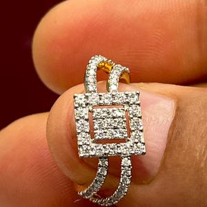 Puede incluir: Un anillo de oro con un centro cuadrado de diamantes y una doble banda. El anillo est&aacute; incrustado con peque&ntilde;os diamantes transparentes. El anillo se sostiene entre dos dedos.