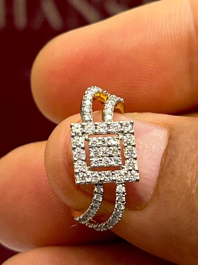 Puede incluir: Un anillo de oro con un centro cuadrado de diamantes y una doble banda. El anillo est&aacute; incrustado con peque&ntilde;os diamantes transparentes. El anillo se sostiene entre dos dedos.