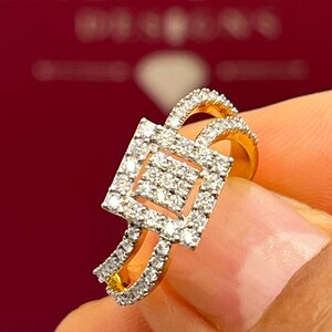 Puede incluir: Un anillo de oro con un centro de racimo de diamantes cuadrados y diamantes m&aacute;s peque&ntilde;os a lo largo de la banda. El anillo tiene un dise&ntilde;o de doble banda y est&aacute; sobre un fondo rojo borroso.