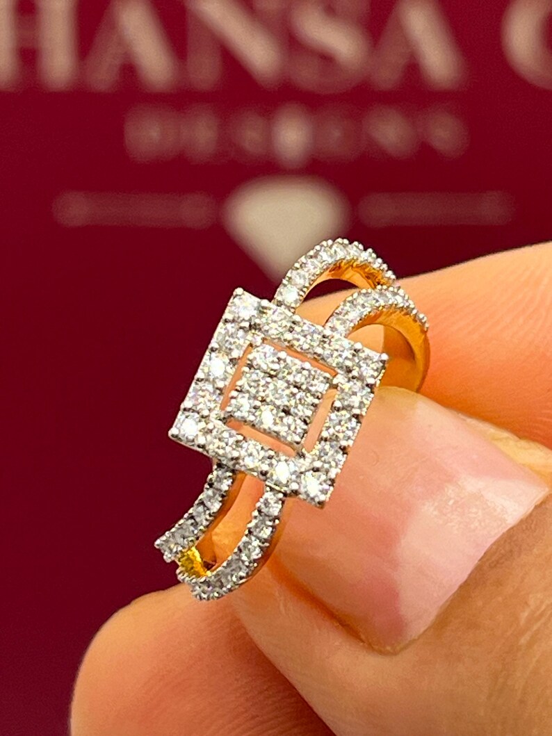Puede incluir: Un anillo de oro con un centro de racimo de diamantes cuadrados y diamantes m&aacute;s peque&ntilde;os a lo largo de la banda. El anillo tiene un dise&ntilde;o de doble banda y est&aacute; sobre un fondo rojo borroso.
