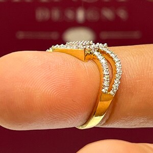 Puede incluir: Un anillo de oro con un dise&ntilde;o de doble banda, incrustado con peque&ntilde;as piedras preciosas transparentes. El anillo est&aacute; sobre un fondo rojo borroso. El anillo est&aacute; en un dedo.
