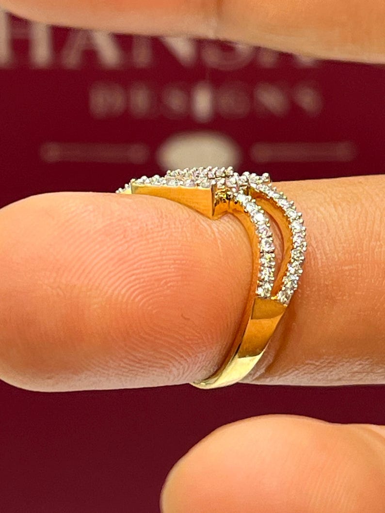 Puede incluir: Un anillo de oro con un dise&ntilde;o de doble banda, incrustado con peque&ntilde;as piedras preciosas transparentes. El anillo est&aacute; sobre un fondo rojo borroso. El anillo est&aacute; en un dedo.