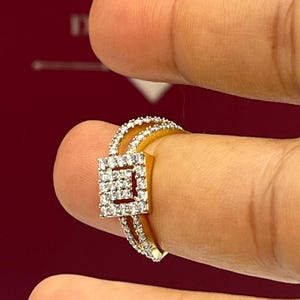 Puede incluir: Un anillo de oro con un centro de racimo de diamantes cuadrados y diamantes m&aacute;s peque&ntilde;os a lo largo de la banda. El anillo se sostiene entre dos dedos sobre un fondo rojo. El dise&ntilde;o del anillo presenta una doble banda.