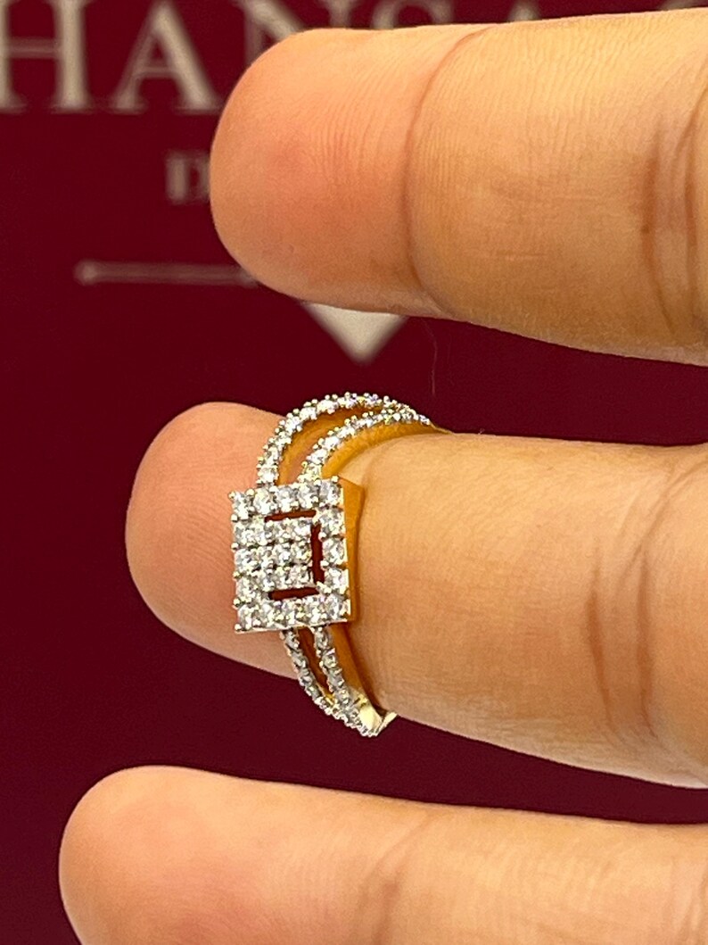 Puede incluir: Un anillo de oro con un centro de racimo de diamantes cuadrados y diamantes m&aacute;s peque&ntilde;os a lo largo de la banda. El anillo se sostiene entre dos dedos sobre un fondo rojo. El dise&ntilde;o del anillo presenta una doble banda.